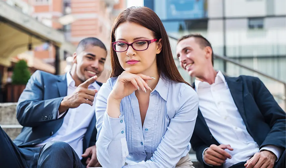 A woman sits apart from a group looking forlorn, whilst a two of her colleagues appear to be talking and laughing at her, set apart in the background.